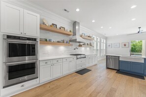Una cocina moderna con armarios blancos, electrodomésticos de acero inoxidable, estantes de madera y vistas al exterior.