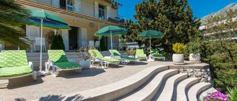 A pool area with lounge chairs and umbrellas, a stone staircase, and a building with balconies.