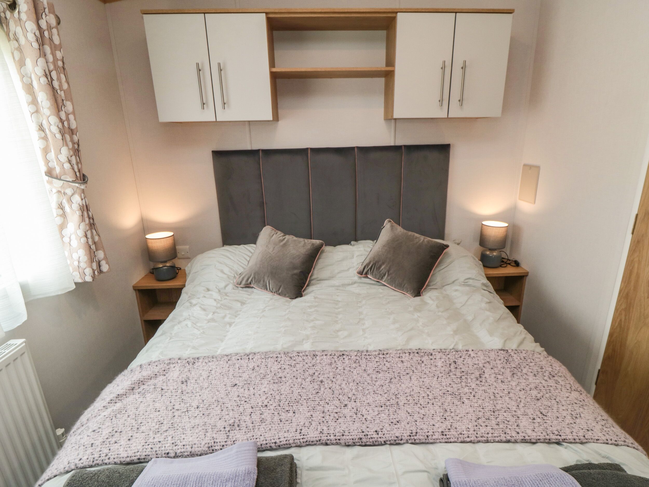 A neatly made bed with a grey headboard, two bedside lamps, and a white wardrobe above.