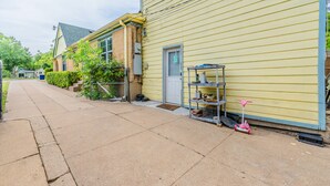 Une rue résidentielle avec une maison jaune, une porte ornée d’une enseigne en forme de « B », une étagère en métal sur laquelle sont disposés divers objets, et un scooter rose.
