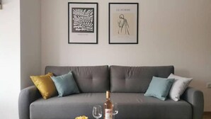 En stue med en grå sofa, to billeder i rammer, en flaske vin og et fad med glas på et træ sofabord.