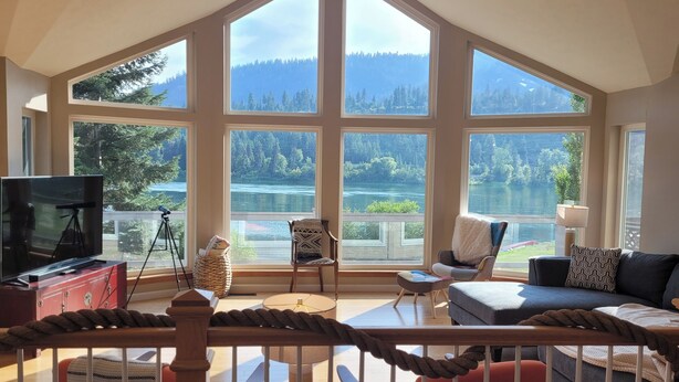 Amplo quarto com grandes janelas que oferecem vista para um lago e uma floresta. Há uma televisão, um tripé e um espaço de estar aconchegante com uma mesa e cadeiras.