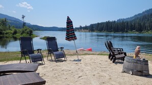 Um cenário à beira do lago com área de estar ao ar livre, incluindo cadeiras e uma mesa, um guarda-sol de praia listrado e um barril de madeira.