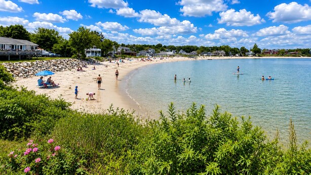 Une plage avec une eau claire, des gens qui se baignent et des maisons en arrière-plan.