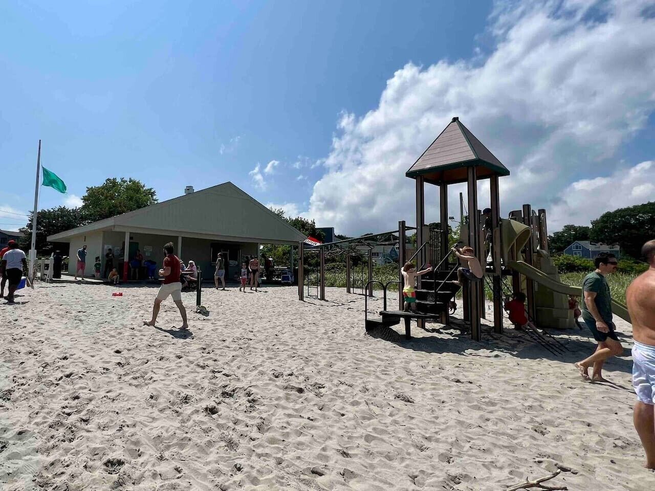 A sandy beach with a playground structure, a building with a green roof, and a flag.