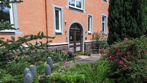 A building with a red facade, a central arched entrance, and a small garden area with potted plants and shrubs.