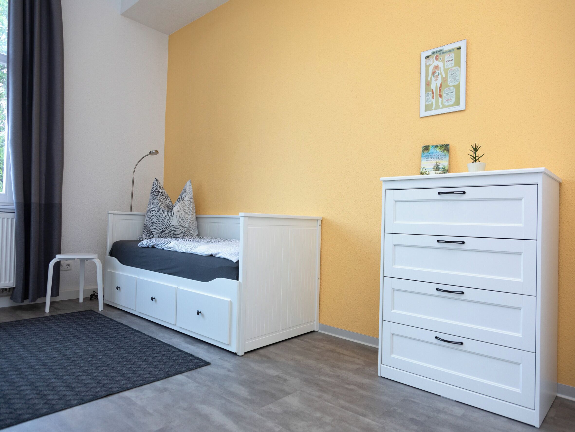 Ein Schlafzimmer mit einem Bett, einer weißen Kommode, einem kleinen Hocker und einem gerahmten Lernplakat an der Wand.