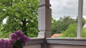 A corner balcony with a round table and two chairs, a vase with purple flowers, and a view of green trees and a building in the background.
