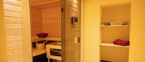 Un sauna aux murs en bois, doté d’une porte en verre et d’une étagère où sont rangées des serviettes et des bouteilles.