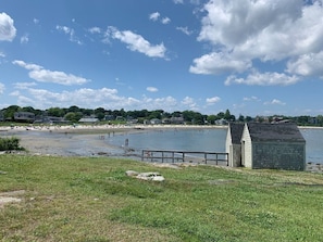 A beach with a small wooden structure, a grassy foreground, and houses in the background.