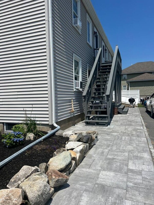 Un edificio residencial con fachada gris, una escalera visible que lleva a la entrada y un camino pavimentado con borde de piedra.