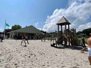 A sandy beach with a playground structure, a building with a green roof, and people walking.