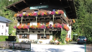 Un chalé tradicional con varios balcones decorados con flores de colores, un letrero que indica que es una pensión, y un coche estacionado adelante.