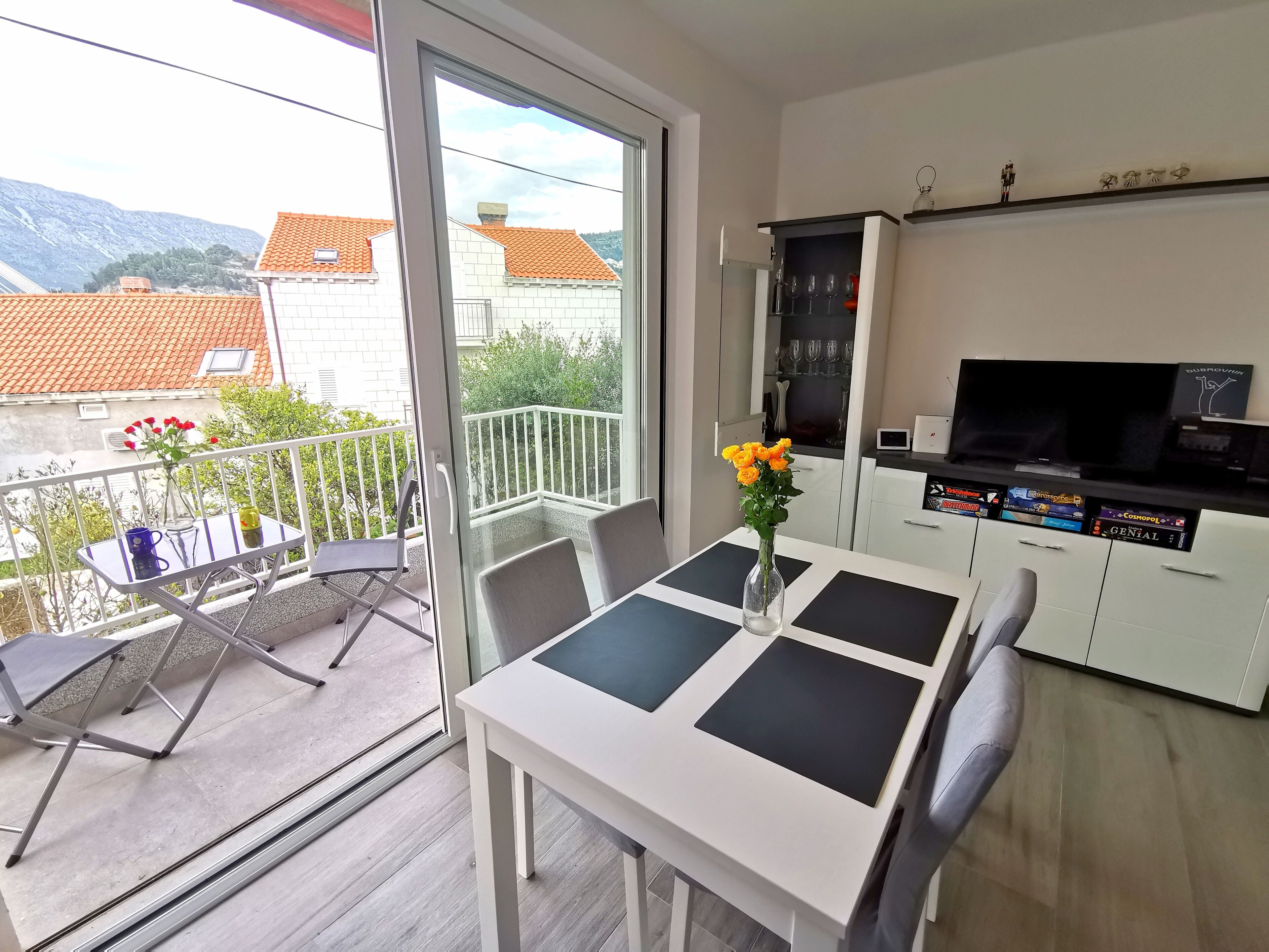 A balcony with a table and chairs, a vase with flowers, and a view of buildings and mountains.