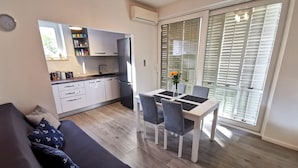 Ein modernes Wohnzimmer mit Sofa, einem Esstisch mit Stühlen, einer Küche mit weißen Schränken und einer Glastür zum Außenbereich.
