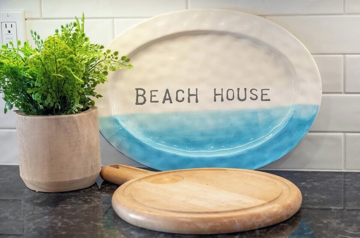 Uma bancada de cozinha com um prato decorativo com a inscrição "BEACH HOUSE", uma tábua de corte de madeira e um vaso com planta.