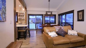 A living room with a sofa, a dining area with chairs, a kitchen area with shelves, and large windows with a view. - Crested Mtn. North L9 (Crested Butte)