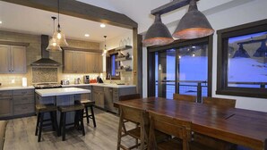 A modern kitchen with wooden cabinets, a large island, pendant lighting, and a view of a snowy landscape through large windows. - Crested Mtn. North L9 (Crested Butte)