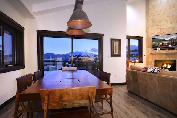 A dining area with a large wooden table, chairs, a pendant light, a fireplace, and a TV mounted on the wall.