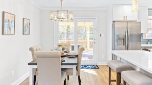 A modern kitchen with white cabinetry, a stainless steel refrigerator, and a dining area with a table and chairs. - Pelican Retreat ~ Screened-in Porch ~ Outdoor Shower ~ Large Back Yard ~ King (Wilmington)
