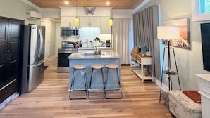 A modern kitchen with wooden flooring, a central island with stools, and pendant lighting. - The Monarch: A Pinehurst Retreat (Carthage)