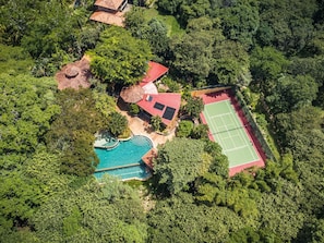 An aerial view of a property with a swimming pool, tennis court, and several buildings surrounded by dense greenery.