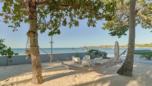 Eine Hängematte mit weißen Stühlen an einem sandigen Strand, umgeben von Bäumen und mit Blick auf das Meer.