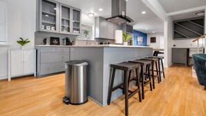A modern kitchen with grey cabinets, a stainless steel trash bin, and wooden stools.