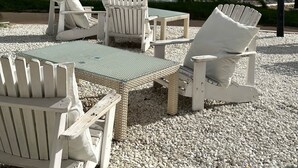 A beachside seating area with white Adirondack chairs and a table, overlooking the ocean with a rocky shoreline and palm trees. - Villa El Barranco (Teulada)