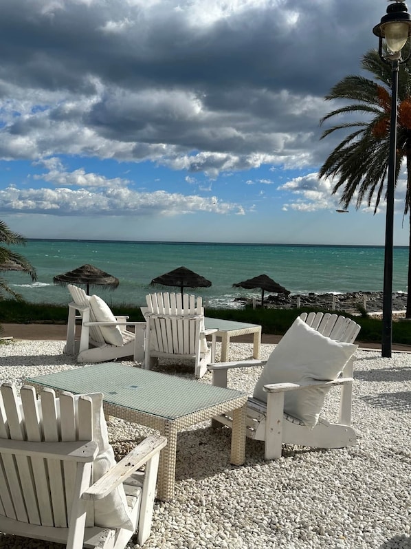 A beachside seating area with white Adirondack chairs and a table, overlooking the ocean with a rocky shoreline and palm trees. - Villa El Barranco (Teulada)