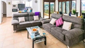 A living room with two grey sofas, a glass coffee table, and a kitchen in the background. - Villa El Barranco (Teulada)