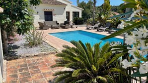 A poolside area with a white building, outdoor furniture, and palm trees. - Villa El Barranco (Teulada)