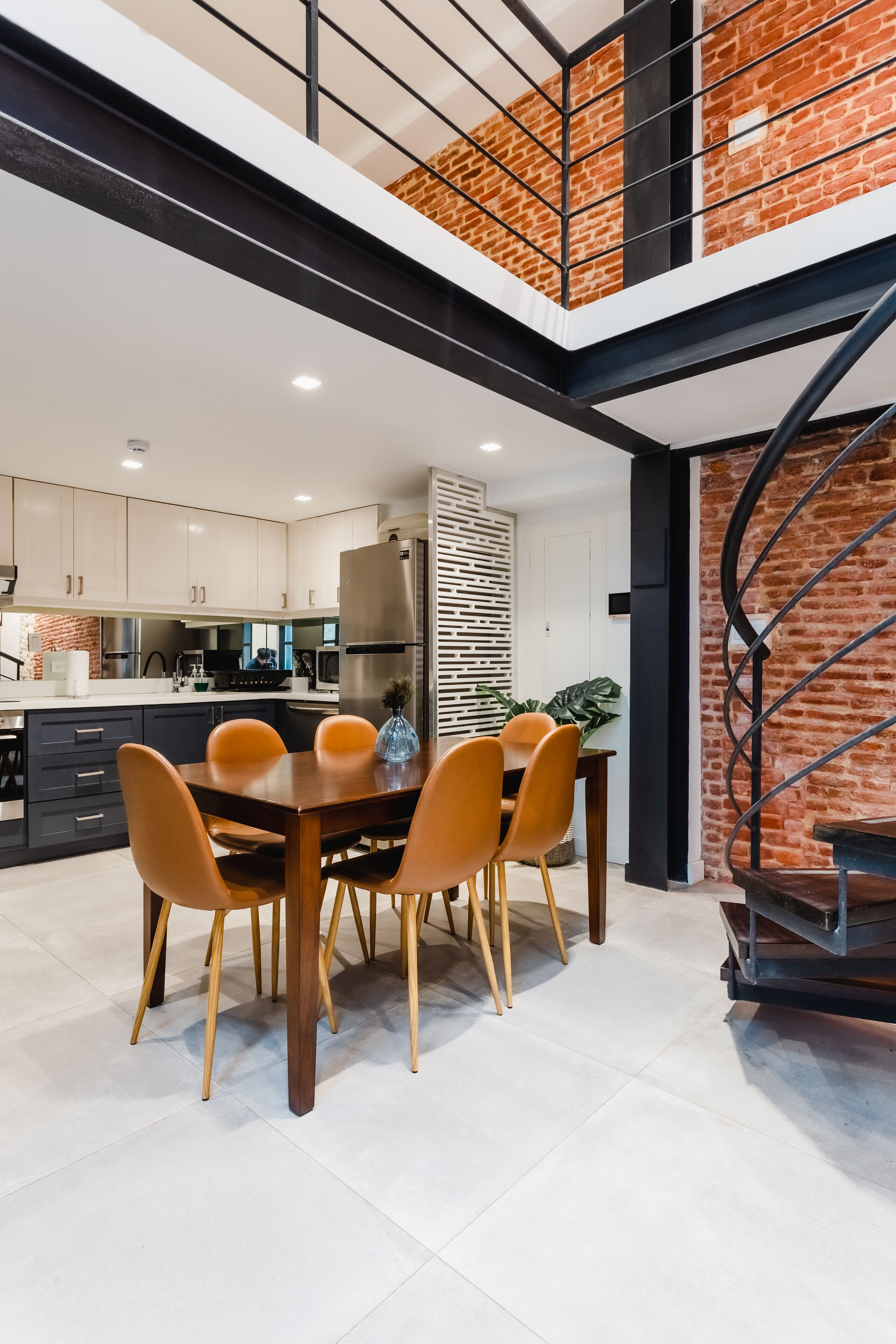 A modern kitchen with white cabinets, a dining area with a wooden table and orange chairs, and a brick wall with a spiral staircase.