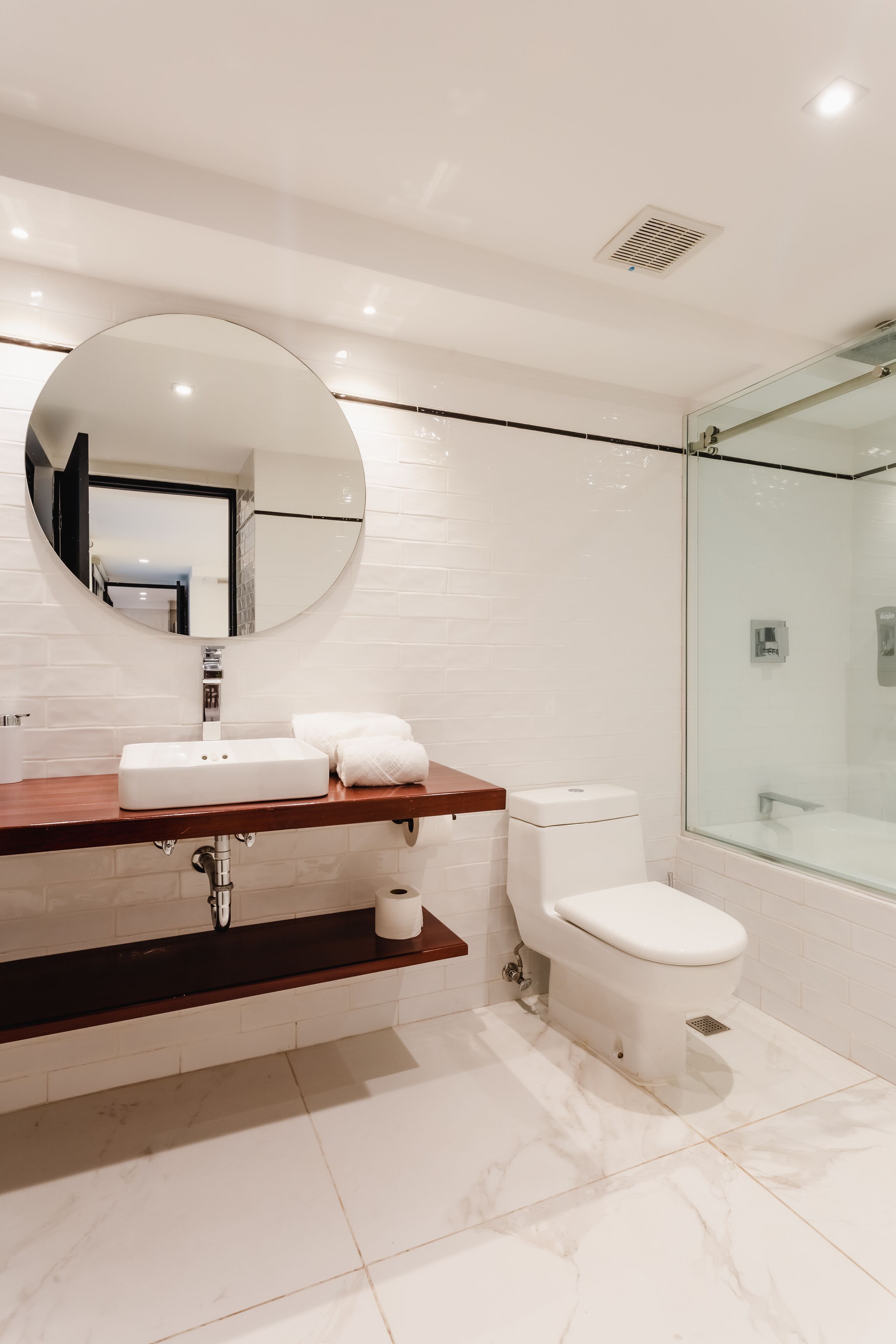 Een moderne badkamer met een witte marmeren vloer, een houten wastafelmeubel, een ronde spiegel, een toilet en een douchecabine van glas.