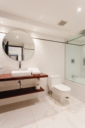 Un baño moderno con piso de mármol blanco, tocador de madera, espejo redondo, inodoro y cabina de ducha de vidrio.