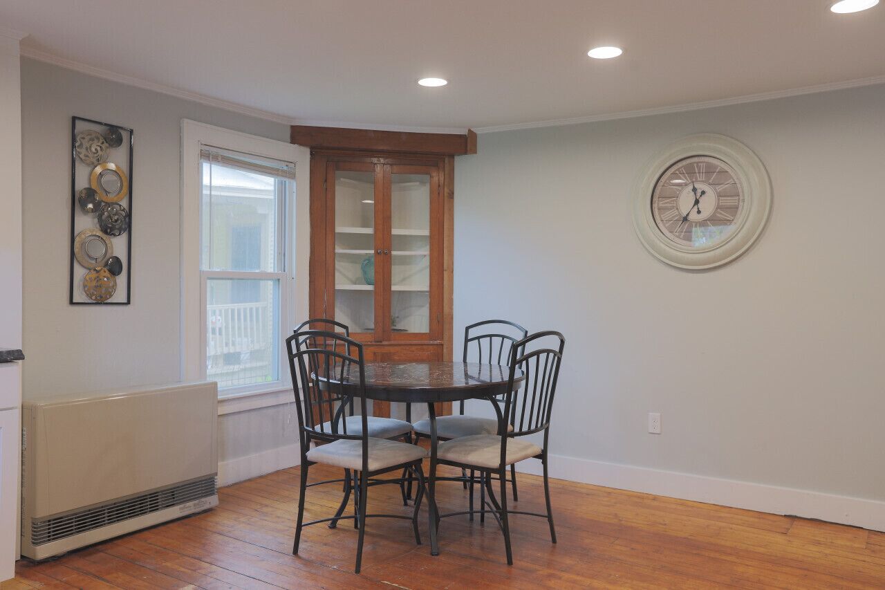 A dining area with a round table and four chairs, a wooden cabinet, a radiator, and a wall clock.