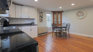 Une cuisine avec des meubles blancs, une cuisinière noire et un coin repas comprenant une table et des chaises en bois.