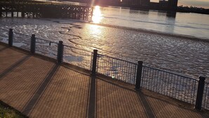 Un paseo marítimo con barandilla de metal, una pasarela de madera y el horizonte de una ciudad al fondo.