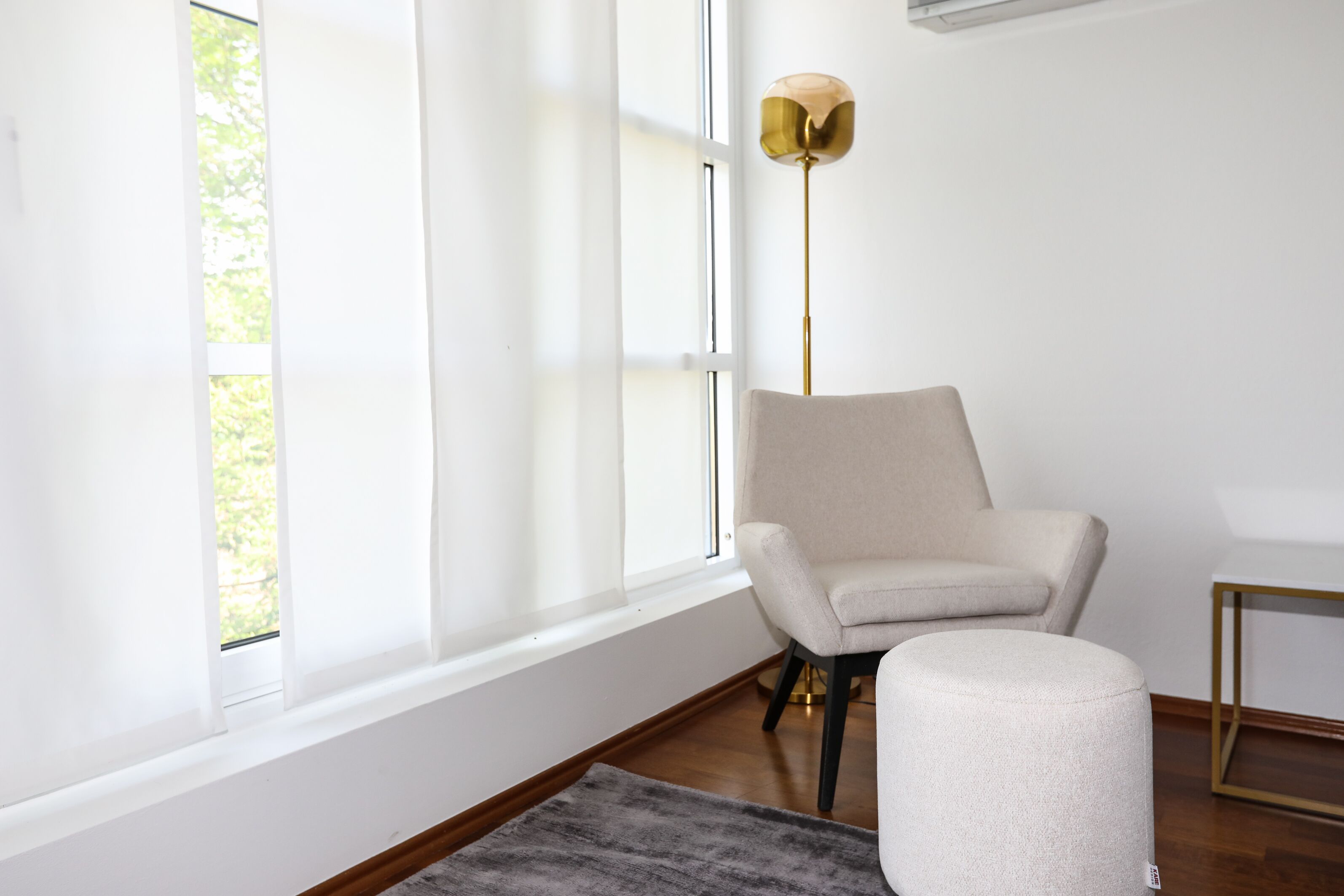 Une pièce avec une grande fenêtre, un fauteuil, un petit pouf et un lampadaire.