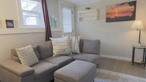 Ein Wohnzimmer mit einem grauen Ecksofa, einem gemusterten Zierkissen, einem kleinen Beistelltisch mit einer Lampe und einem gerahmten Bild an der Wand.