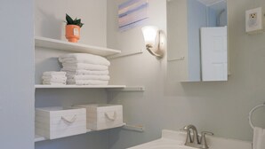 Ein Badezimmer mit weißen Regalen, auf denen Handtücher und ein Topf mit einer Pflanze stehen, einer Wandleuchte, einer weißen Tür und einem Waschbecken mit Armatur.