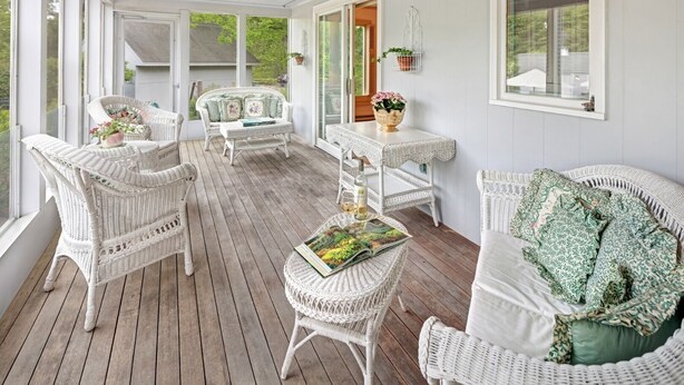 En inglasad veranda med möbler i rotting, ett bord med en vas med blommor och utsikt över omgivningen.