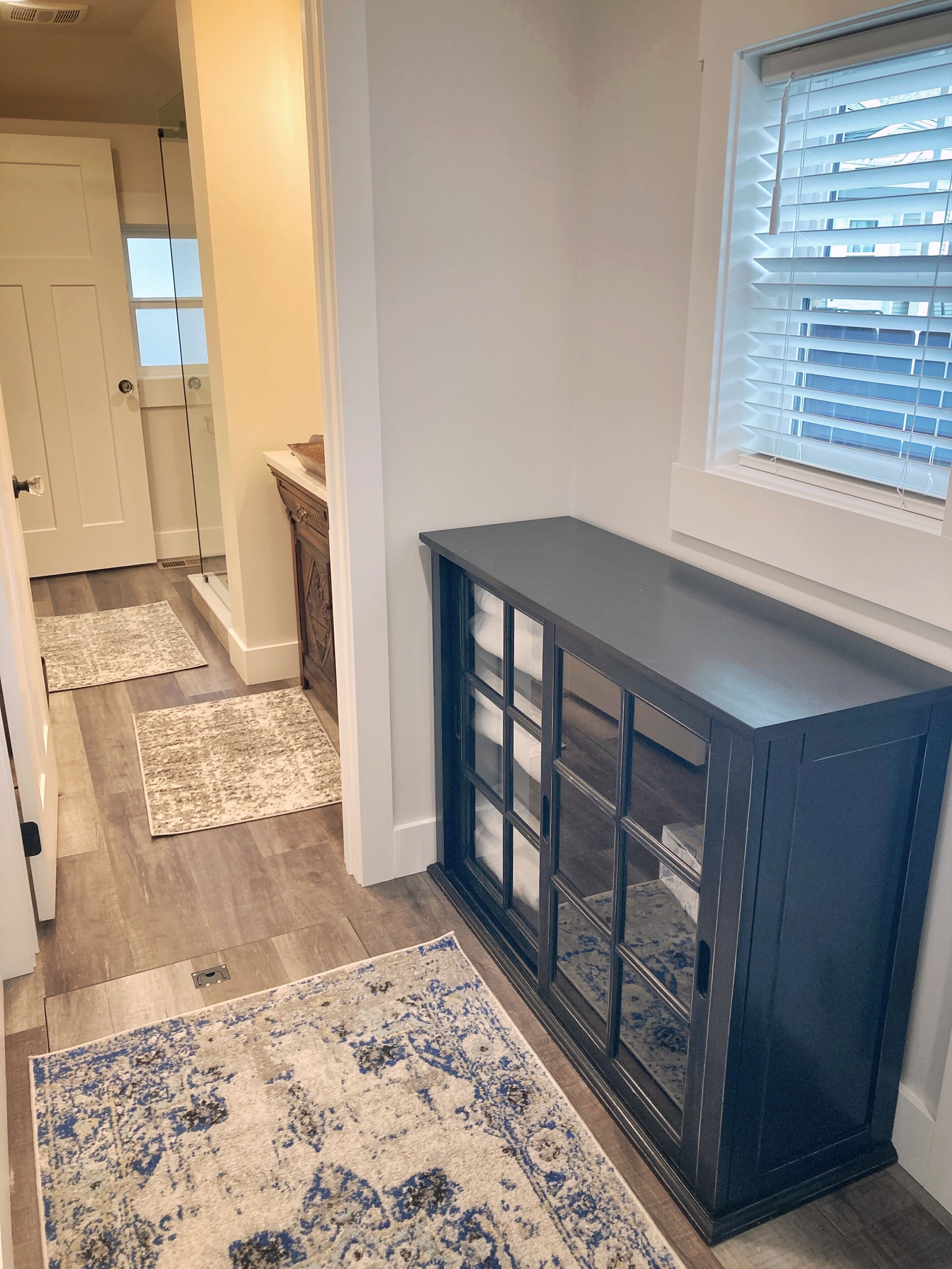 A hallway with a dark wooden console table, a window with blinds, and a door leading to a bathroom.