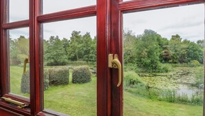 A view through a window with a wooden frame and a brass handle, overlooking a pond surrounded by lush greenery and trees. - 10 Ruston Reaches (Norwich)