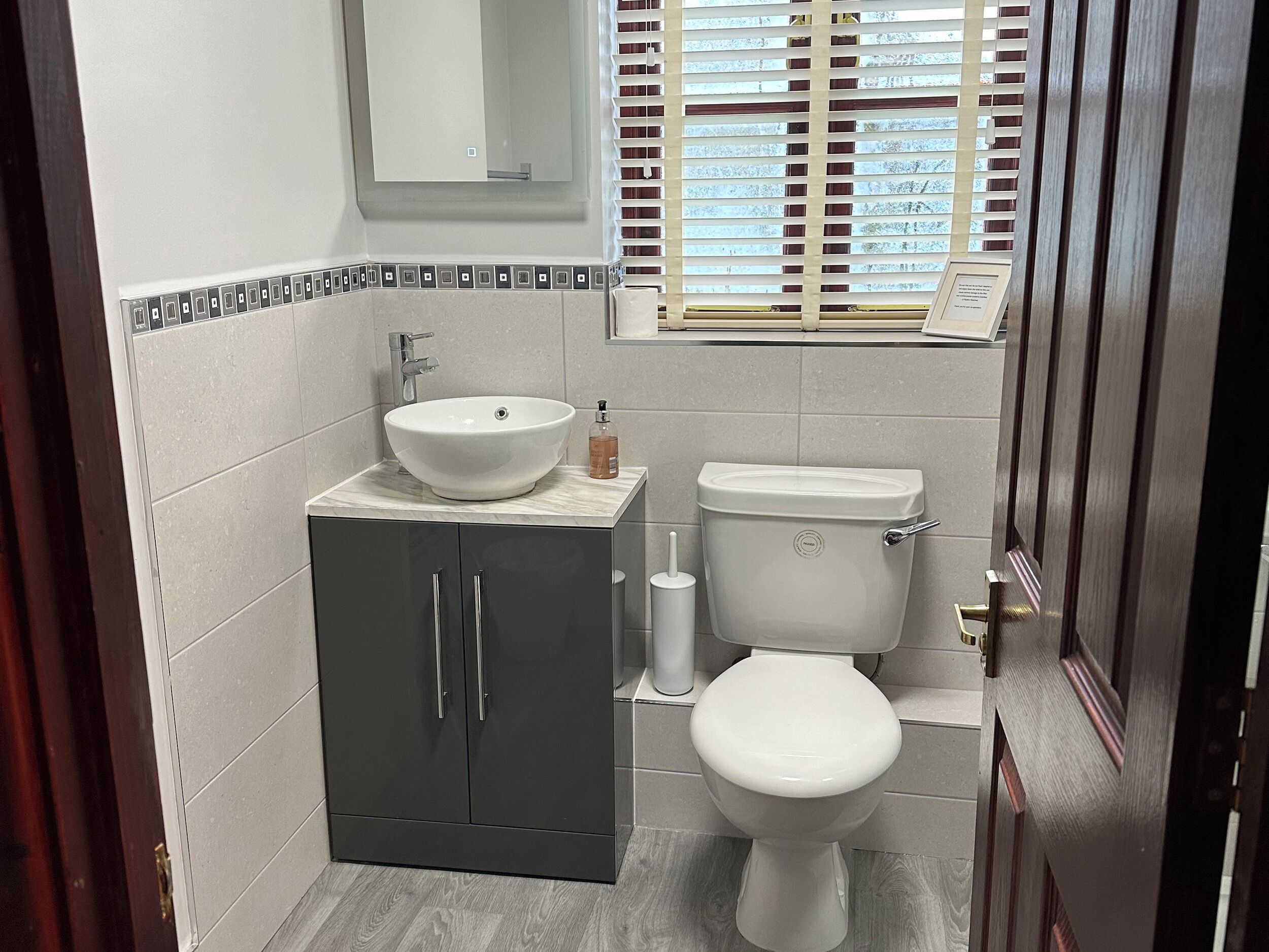 A bathroom with a white sink, dark cabinets, a toilet, and a window with blinds.