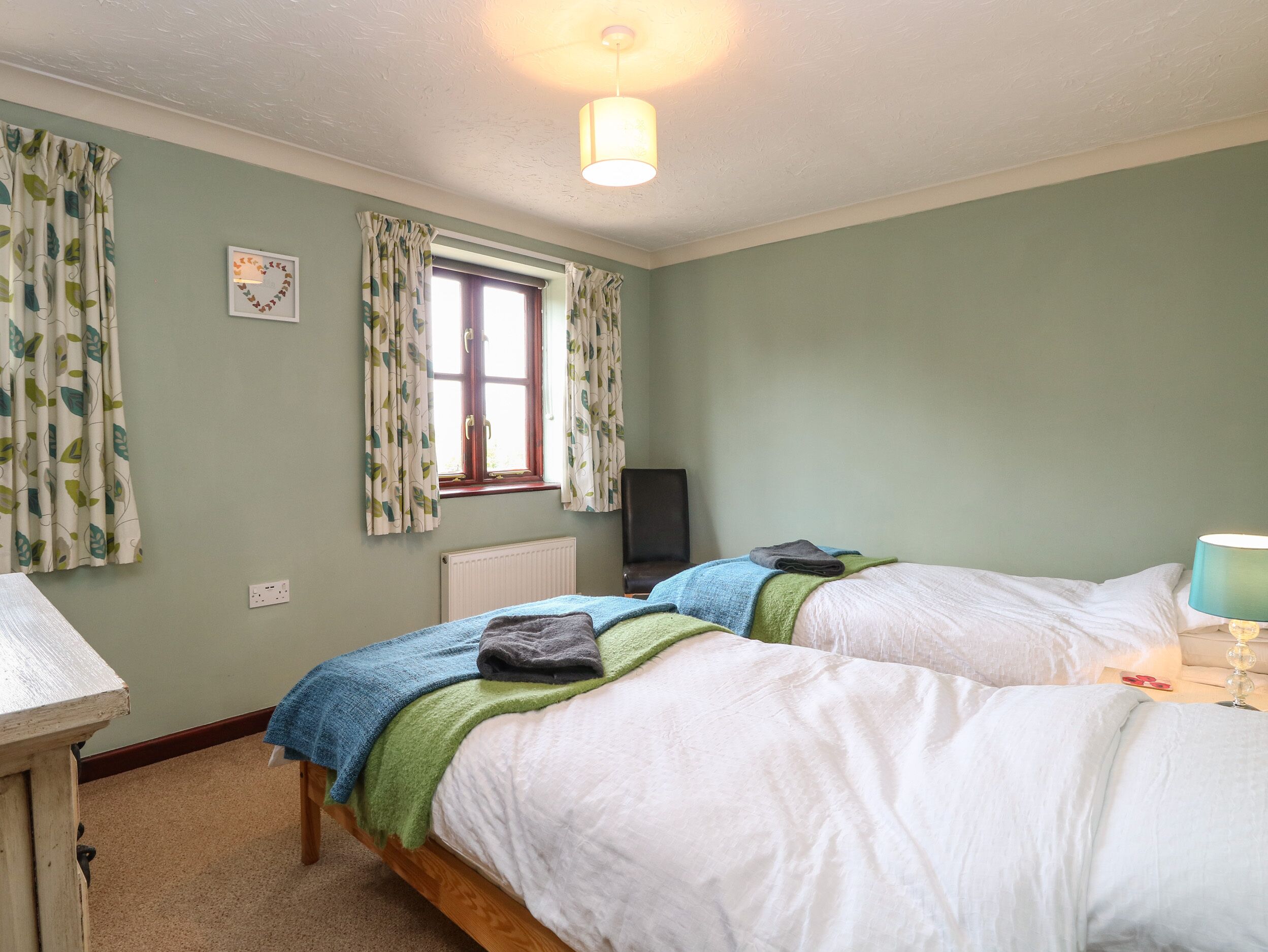 A bedroom with two beds, a window with curtains, a radiator, a desk, and a lamp.