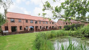 A brick building with multiple windows and doors, a grassy lawn, and a pond with reeds. - 10 Ruston Reaches (Norwich)