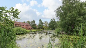 A serene riverside view with a red brick building, lush greenery, and a calm water surface with lily pads. - 10 Ruston Reaches (Norwich)