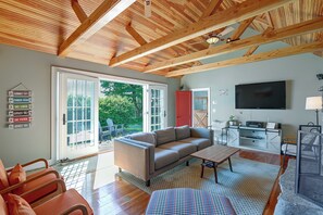 A spacious living area with a wooden ceiling, a sofa, a coffee table, and a television. There is a sliding glass door leading to an outdoor area with greenery.