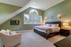 A bedroom with a sloped ceiling, a large window, a bed, a chair, and a bedside table with lamps.